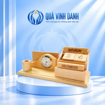 Minh họa bộ để bàn gỗ có ống bút tròn, đồng hồ viền màu vàng và lịch hai khối số giữ SATURDAY 24 DECEMBER trên bục Quà Vinh Danh
