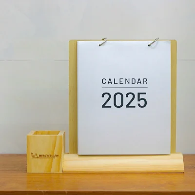 Ảnh thật toàn cảnh lịch đế gỗ vát, bìa trắng CALENDAR 2025 và ống bút vuông mang logo Michelin ở bên trái