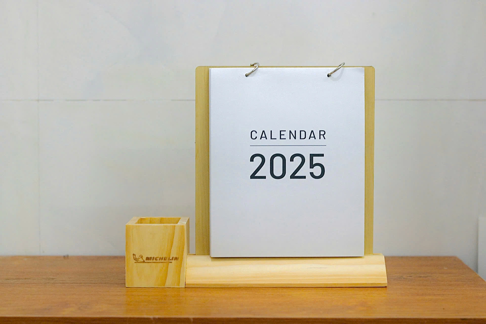Ảnh thật toàn cảnh lịch đế gỗ vát, bìa trắng CALENDAR 2025 và ống bút vuông mang logo Michelin ở bên trái