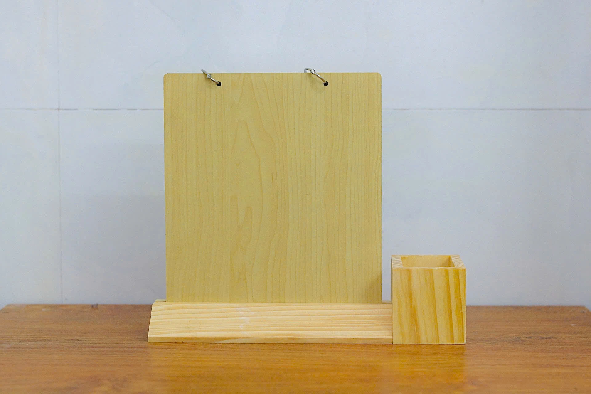 Ảnh thật mặt sau bộ lịch với tấm đỡ gỗ trống, hai khoen ở trên, đế vát và ống bút bên phải do đổi hướng nhìn
