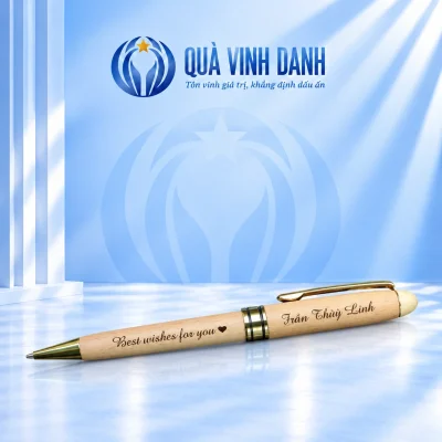 Bút gỗ sáng BGV 001 với đầu và cài màu vàng, giữ lời chúc cùng tên Trần Thùy Linh trên nền Quà Vinh Danh