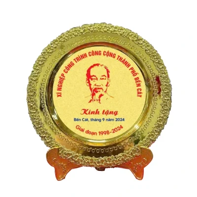 Ảnh thật đĩa mạ vàng có chân dung, tên Xí nghiệp Công trình Công cộng thành phố Bến Cát và giai đoạn 1998–2024, giữ nguyên chữ nguồn