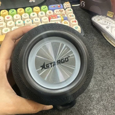 Ảnh chụp nguồn loa StarGO Boom Lite đen cầm trên tay thấy mặt bạc logo hãng bề mặt dạng lưới và hộp Boom Lite phía sau
