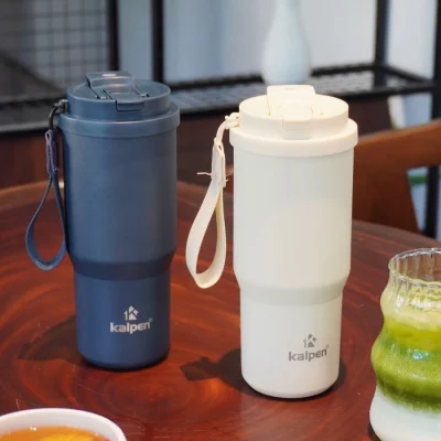 Ảnh thật nguồn hai ly Kalpen DA05 xanh than và trắng kem trên bàn gỗ giữ nguyên logo dây đeo nắp và bối cảnh