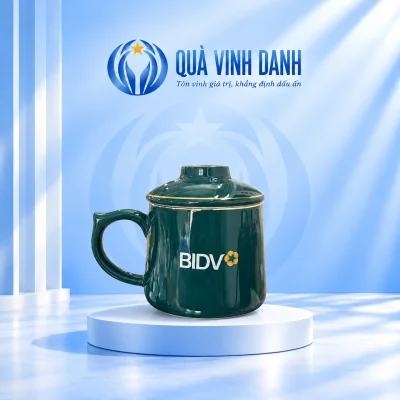 Minh họa ly lọc trà xanh đậm nắp núm tròn, quai trái và chữ BIDV cùng biểu trưng nguồn trên bục Quà Vinh Danh